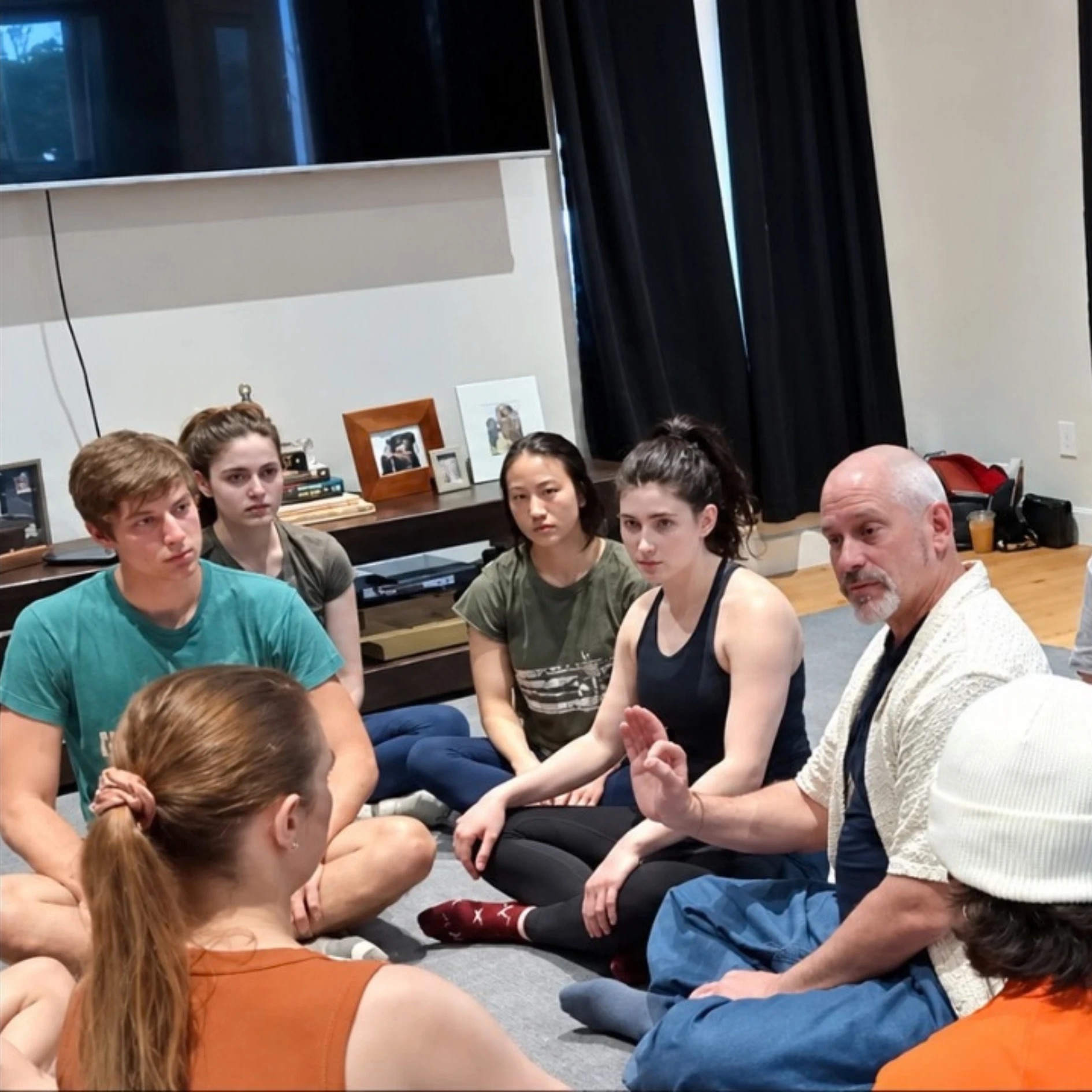Workshop group seated in a circle on a studio floor, listening to the instructor speak and gesture